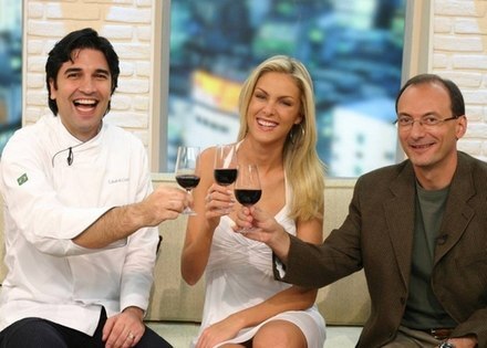 Edu Guedes, Ana Hickmann e Britto Júnior, apresentadores da primeira fase do Hoje em Dia em 2007, sentados lado a lado em um sofá, brindando com taças de vinho tinto, diante de parede de tijolos e janela com vista urbana desfocada ao fundo