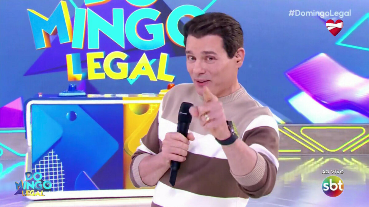Celso Portiolli, apresentador do Domingo Legal no SBT, segura microfone e aponta. No canto superior direito, um coração vermelho aparece na tela
