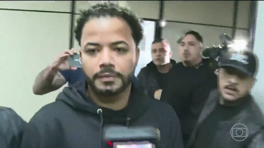 Influenciador Hytalo Santos em um ambiente interno. Ele está no centro da imagem, olhando para a câmera com uma expressão séria e usando um moletom preto. Ao seu redor, outras pessoas aparecem desfocadas, com alguns segurando celulares e câmeras com flashes. O logo da TV Globo é visível no canto inferior direito.