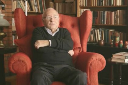 Luis Fernando Verissimo sentado em uma ampla poltrona vermelha, cercado por estante repleta de livros e itens decorativos, incluindo bonecas russas, em ambiente que remete a um escritório ou biblioteca. Cena exibida pelo *Fala Brasil* (Record) na cobertura sobre sua internação em Porto Alegre