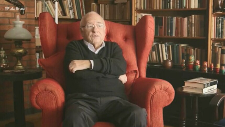 Luis Fernando Verissimo sentado em uma ampla poltrona vermelha, cercado por estante repleta de livros e itens decorativos, incluindo bonecas russas, em ambiente que remete a um escritório ou biblioteca. Cena exibida pelo *Fala Brasil* (Record) na cobertura sobre sua internação em Porto Alegre