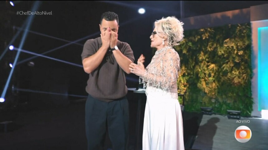 Luiz Lira, emocionado, cobre o rosto com as mãos ao ser anunciado campeão da primeira temporada do reality culinário Chef de Alto Nível, ao lado de Ana Maria Braga no palco da final ao vivo