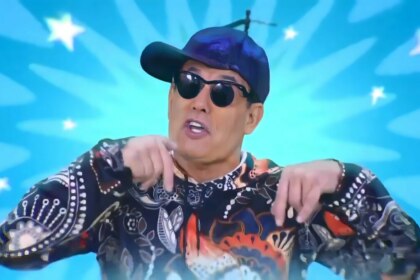 Sérgio Mallandro no comercial do Porta dos Desesperados, usando boné azul e camisa colorida diante de fundo azul vibrante