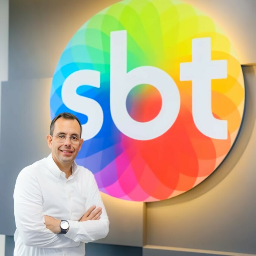 O executivo Vicente Varela posa sorrindo, com os braços cruzados, em frente ao logo colorido do SBT.