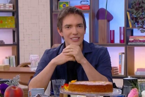 O jornalista Alessandro Jodar sorri na mesa do Mais Você, que está repleta de comida e um bolo. Ele usa uma camisa azul com uma camiseta preta.