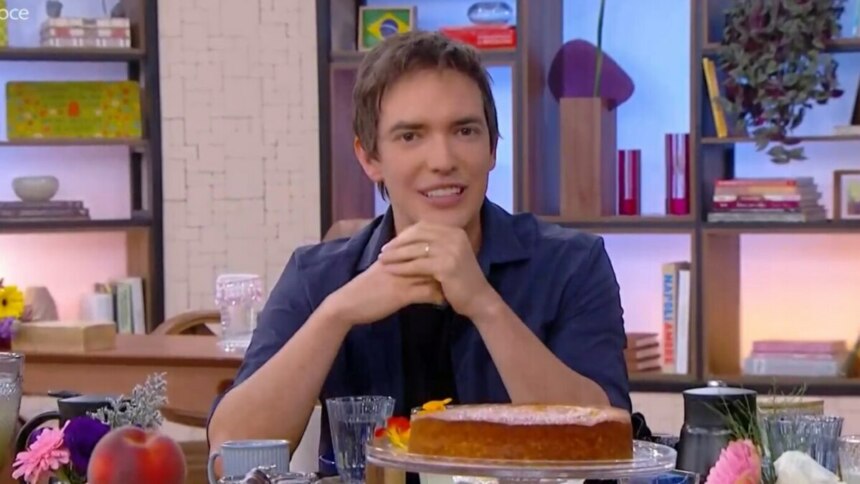 O jornalista Alessandro Jodar sorri na mesa do Mais Você, que está repleta de comida e um bolo. Ele usa uma camisa azul com uma camiseta preta.