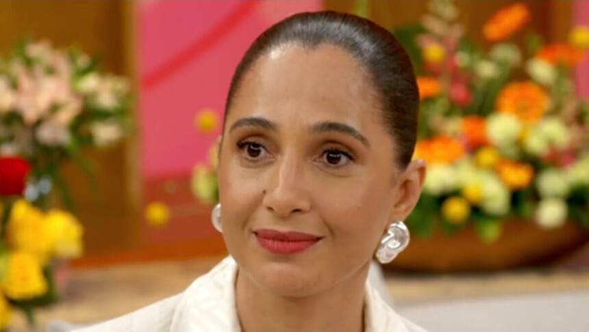 A atriz Camila Pitanga, com o cabelo preso em um coque, usa grandes brincos brancos. Ela está sorrindo discretamente em uma participação no programa Encontro, com um fundo de flores borrado.