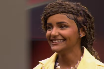 Camille Vitória, de cabelos trançados, sorrindo e olhando para o lado, com uma jaqueta amarela em trecho da Prova do Jingle do programa Estrela da Casa.