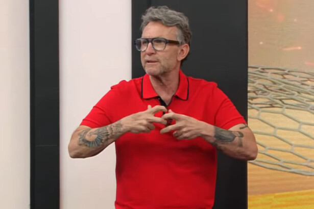 O apresentador e comentarista Craque Neto, de barba e cabelo grisalhos, usa óculos e uma camisa polo vermelha. Ele está gesticulando com as mãos no estúdio do programa Apito Final, com um fundo preto e um gráfico digital abstrato ao seu lado.