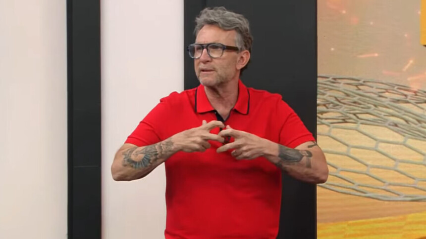 O apresentador e comentarista Craque Neto, de barba e cabelo grisalhos, usa óculos e uma camisa polo vermelha. Ele está gesticulando com as mãos no estúdio do programa Apito Final, com um fundo preto e um gráfico digital abstrato ao seu lado.