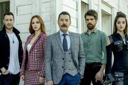 Os atores da novela Cruel Istambul (Fikret Kuskan, Deniz Ugur, Berker Guven, Mine Tugay e Sera Kutlubey) estão posando para a foto, sérios e alinhados, em frente a uma casa branca.