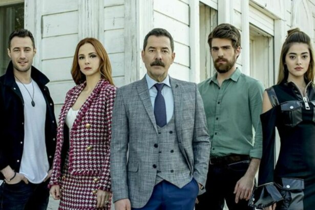 Os atores da novela Cruel Istambul (Fikret Kuskan, Deniz Ugur, Berker Guven, Mine Tugay e Sera Kutlubey) estão posando para a foto, sérios e alinhados, em frente a uma casa branca.
