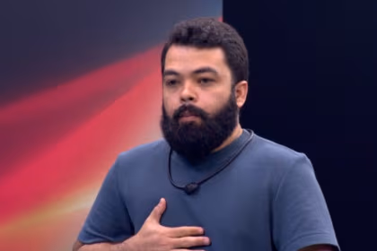 O participante Daniel Sobral, barbudo, com uma camisa azul, gesticulando no peito, no cenário da Prova do Jingle do reality show Estrela da Casa.