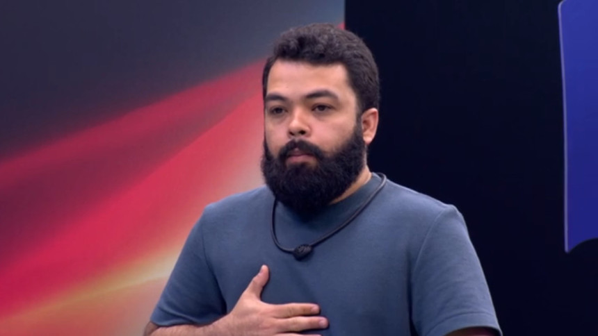 O participante Daniel Sobral, barbudo, com uma camisa azul, gesticulando no peito, no cenário da Prova do Jingle do reality show Estrela da Casa.