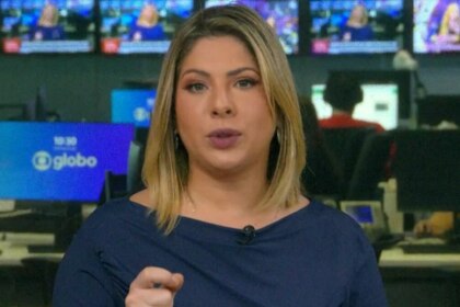 Daniela Lima com cabelos loiros e curtos, usando uma blusa azul-marinho, olhando para a câmera e gesticulando com a mão direita no estúdio da GloboNews, com monitores ao fundo.