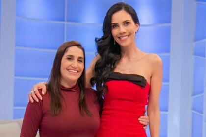 Daniele Hypolito e Daniela Albuquerque posando para a foto. Daniele, de blusa vinho, abraça Daniela, que usa um vestido vermelho tomara que caia com um laço preto na frente, no cenário do programa Sensacional.