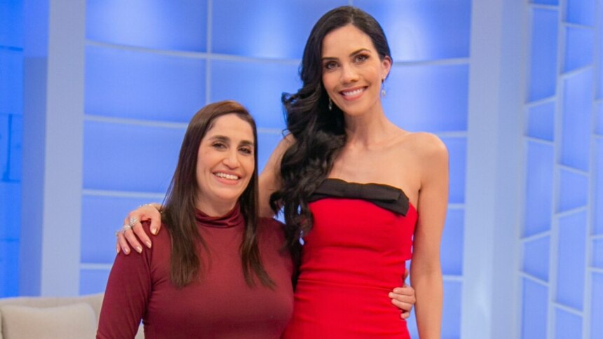 Daniele Hypolito e Daniela Albuquerque posando para a foto. Daniele, de blusa vinho, abraça Daniela, que usa um vestido vermelho tomara que caia com um laço preto na frente, no cenário do programa Sensacional.