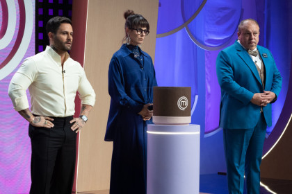 Os jurados do MasterChef Confeitaria, Diego Lozano, Helena Rizzo e Erick Jacquin, posam sérios ao lado de um objeto cilíndrico marrom, no estúdio do programa.