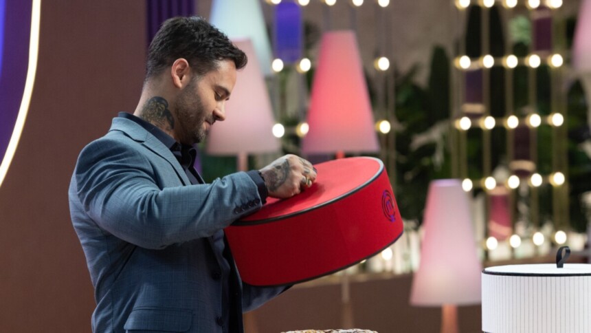 O chef Diego Lozano, de terno azul-claro com detalhes e braços tatuados, inclina-se sobre um bolo em uma caixa vermelha em um cenário iluminado de MasterChef Confeitaria.