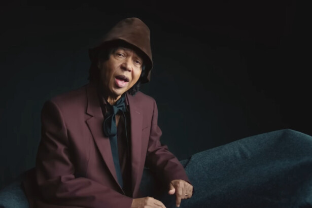 Djavan de terno vinho, gravata preta, sentado em um cenário escuro e cantando Um Brinde