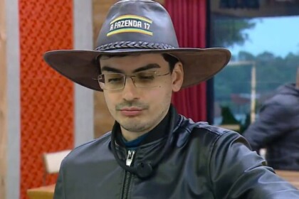 Dudu Camargo usa o chapéu de Fazendeiro marrom de A Fazenda 17. Ele veste jaqueta de couro preta, óculos e olha para baixo.