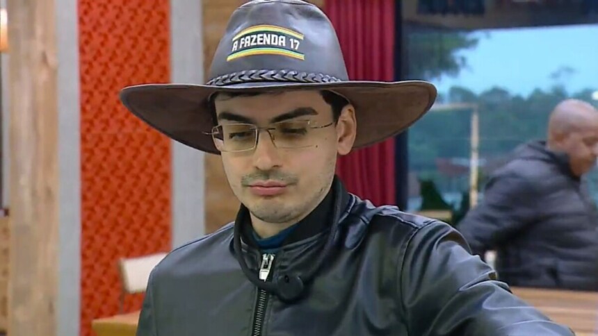 Dudu Camargo usa o chapéu de Fazendeiro marrom de A Fazenda 17. Ele veste jaqueta de couro preta, óculos e olha para baixo.