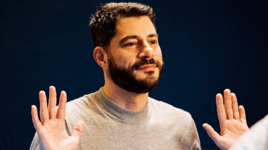 O jornalista Evaristo Costa, com barba e bigode, está com as mãos levantadas à altura dos ombros, com as palmas voltadas para a frente, em um gesto de "pare". Ele usa um suéter cinza-claro e olha para o lado.