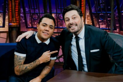 Felipe Araújo aponta para Danilo Gentili no cenário do The Noite, um retrato da dupla em um talk show.