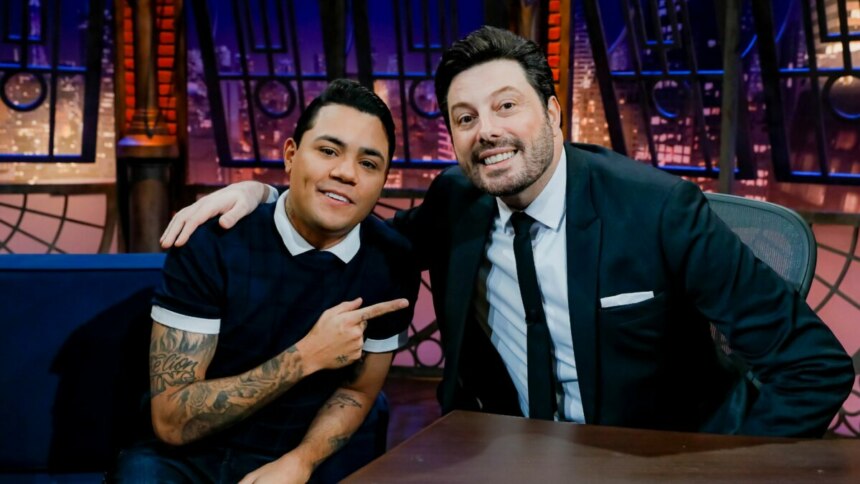 Felipe Araújo aponta para Danilo Gentili no cenário do The Noite, um retrato da dupla em um talk show.