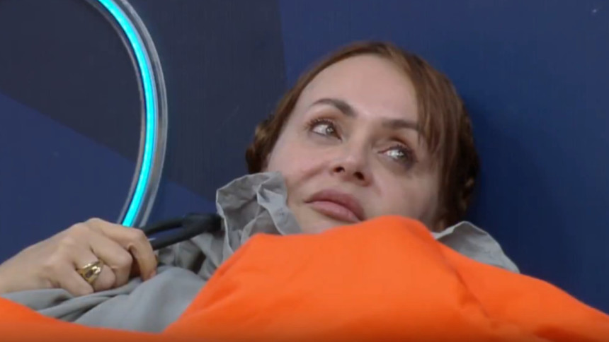 A atriz Gabriela Spanic deitada, com a cabeça apoiada em um travesseiro e coberta por uma manta laranja em A Fazenda 17. Ela parece pensativa ou triste.