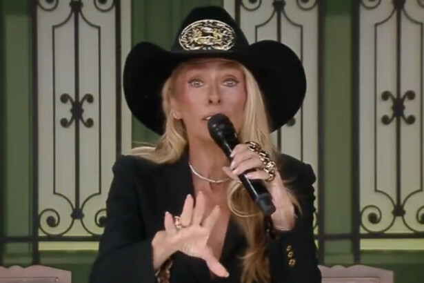 A apresentadora Adriane Galisteu, de terno preto e chapéu de caubói, segura um microfone e gesticula com a mão, em uma cena do reality show A Fazenda.