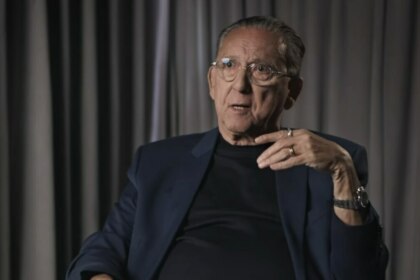 Galvão Bueno em entrevista, gesticulando com expressão séria, diante de cortinas em cenário intimista e iluminado.