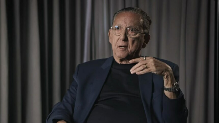 Galvão Bueno em entrevista, gesticulando com expressão séria, diante de cortinas em cenário intimista e iluminado.