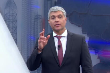 Joel Datena, com cabelos e barba grisalhos, usa um terno escuro, camisa branca e gravata vermelha. Ele está no estúdio do programa Brasil Urgente, olhando diretamente para a câmera e gesticulando com a mão direita.