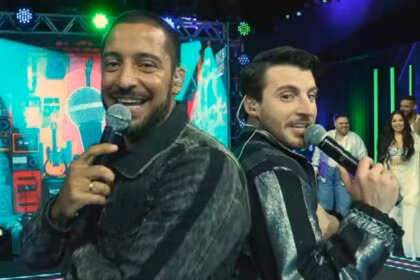 Juceir Jr e Gabriel Smaniotto, participantes do Estrela da Casa, posam sorrindo de costas um para o outro, segurando microfones no palco.