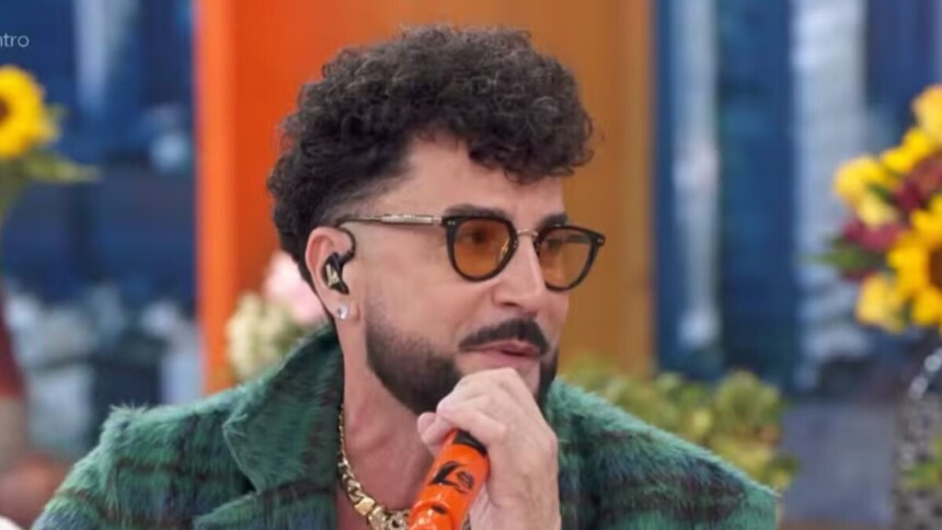 O cantor Latino, usando uma jaqueta verde peluda e óculos escuros com armação amarela, está segurando um microfone laranja com seu nome e sorrindo em uma entrevista no Encontro.