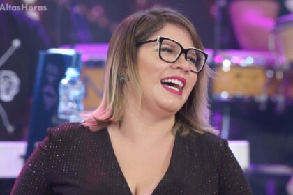 A cantora Marília Mendonça sorri e canta no programa Altas Horas. Ela usa óculos grandes de aro preto, batom vermelho e um vestido preto brilhante.