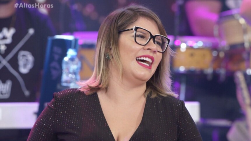 A cantora Marília Mendonça sorri e canta no programa Altas Horas. Ela usa óculos grandes de aro preto, batom vermelho e um vestido preto brilhante.