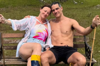 Mateus Solano sem camisa e Paula Braun com shorts jeans sorriem para a câmera, abraçados em um banco de madeira em meio à natureza.