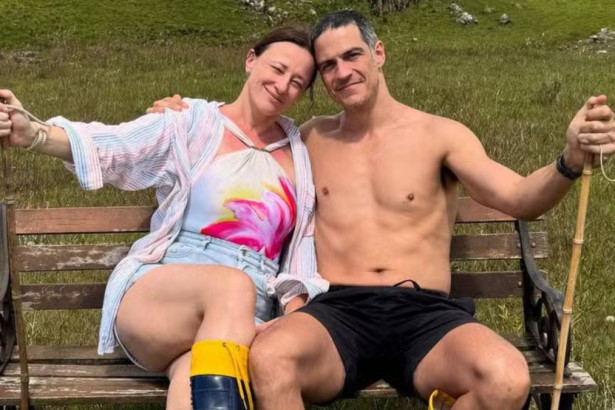 Mateus Solano sem camisa e Paula Braun com shorts jeans sorriem para a câmera, abraçados em um banco de madeira em meio à natureza.