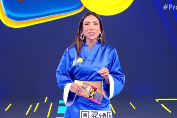 A apresentadora Patricia Abravanel está no palco do programa Silvio Santos. Ela veste um quimono azul com um cinto dourado e segura um bloco da Tele Sena de Primavera. Patrícia sorri e olha para a câmera.