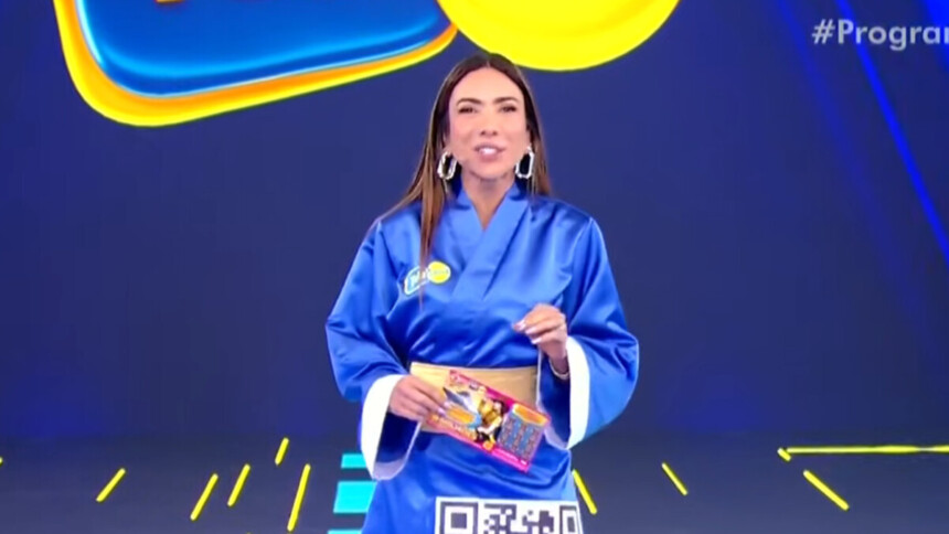 A apresentadora Patricia Abravanel está no palco do programa Silvio Santos. Ela veste um quimono azul com um cinto dourado e segura um bloco da Tele Sena de Primavera. Patrícia sorri e olha para a câmera.