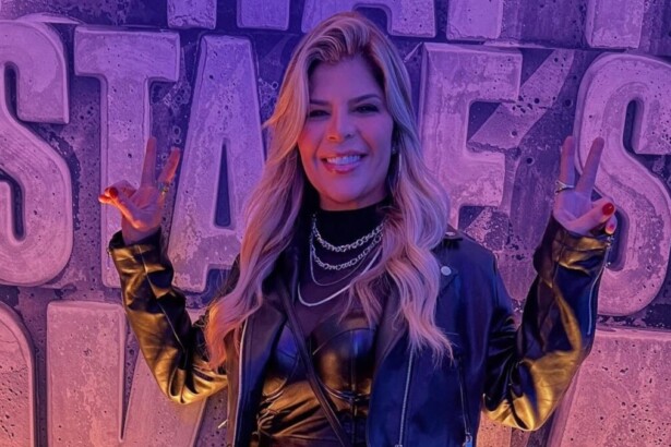 A cantora Paula Mattos sorri, fazendo um sinal de paz com cada mão. Ela usa jaqueta de couro preta, colares, e o fundo é roxo com grandes letras em 3D.