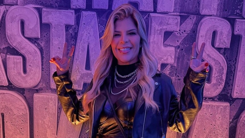 A cantora Paula Mattos sorri, fazendo um sinal de paz com cada mão. Ela usa jaqueta de couro preta, colares, e o fundo é roxo com grandes letras em 3D.