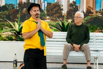O comediante Saideira, ajoelhado, com as mãos unidas e uma garrafa de cachaça ao lado, e Carlos Alberto de Nóbrega sentados em um banco, em trecho de A Praça é Nossa