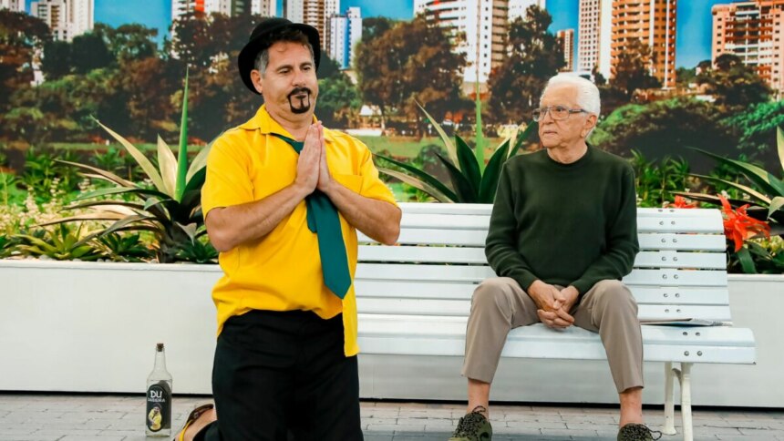 O comediante Saideira, ajoelhado, com as mãos unidas e uma garrafa de cachaça ao lado, e Carlos Alberto de Nóbrega sentados em um banco, em trecho de A Praça é Nossa