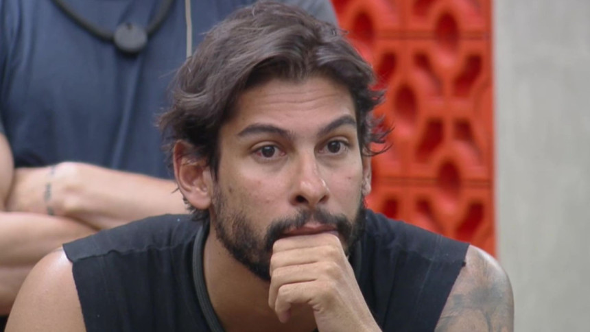 O participante Shia de blusa preta, com barba e cabelos longos, olha para o lado com uma expressão séria, em uma cena do reality show A Fazenda.
