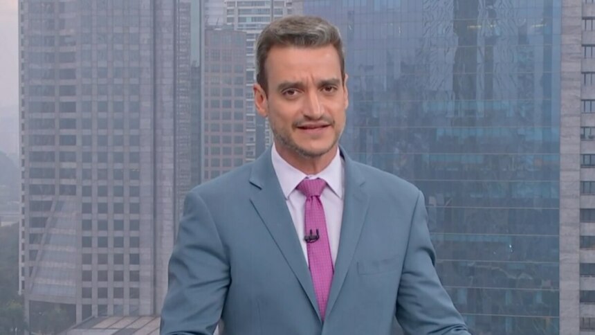 O jornalista Tiago Scheuer está em um estúdio de TV no Bom Dia SP, vestido com um terno azul claro, camisa branca e gravata rosa. O fundo é um cenário de cidade.