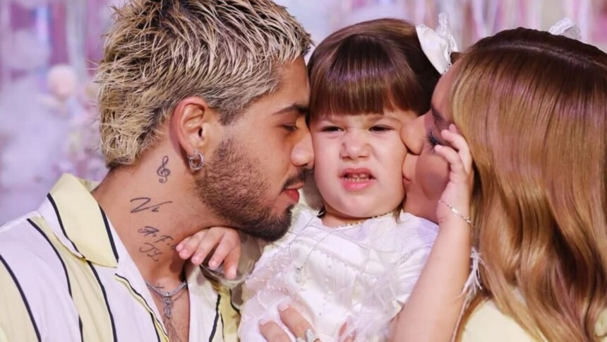 O cantor Zé Felipe, com cabelos descoloridos, beija a cabeça da filha Maria Flor, que está com uma expressão engraçada. A influenciadora Virginia Fonseca beija o rosto da filha ao lado deles em uma foto de família.