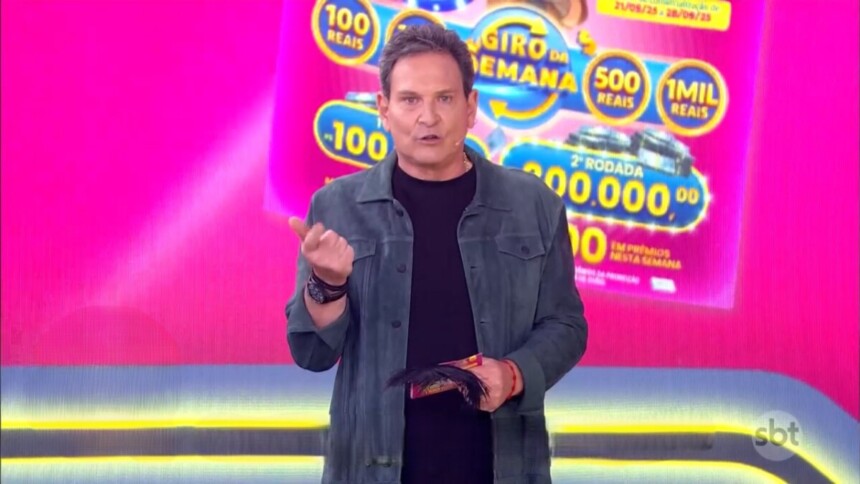 Luis Ricardo no estúdio rosa apresentando a edição 71 da Tele Sena Semanal. Ele segura o bilhete da edição
